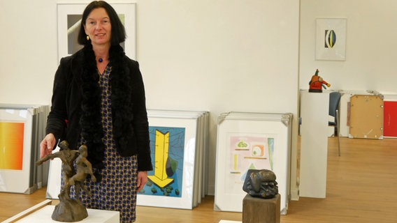 Sabine Isensee, Leiterin der Artothek Oldenburg, steht in einem Raum mit an die Wand gelehnten Bildern im Hintergrund und Skulpturen im Vordergrund. © NDR Foto: Helgard Füchsel