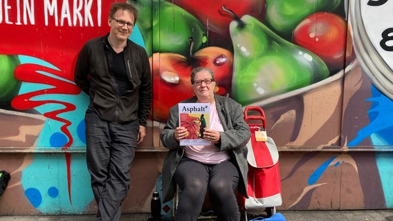 Tina Maschke, Verkäuferin des Straßenmagazins Asphalt, posiert mit einer Ausgabe in der Hand neben Chefredakteur Volker Macke vor einer bunten Wand. © NDR Foto: Andrea Schwyzer