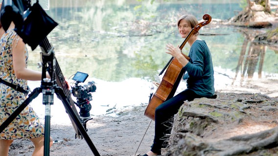 Im Vordergrund eine aufgebaute Kamera, im Hintergrund sitzt Tanja Tetzlaff auf einem Baumstamm und spielt Cello. © Michael Bessert 