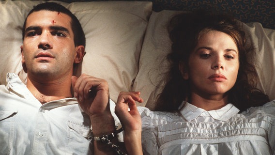 Filmstill "Fessle mich" (1990): Schauspieler Antonio Banderas und Victoria Abril liegen mit Handschellen aneinander gefesselt nebeneinander im Bett © Filmfest München 
