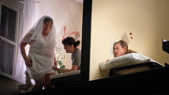 In zwei durch eine Wand getrennte Zimmer sieht man drei Frauen - eine lauscht an der Wand, was die anderen im anderen Zimmer machen. © Thomas Aurin 