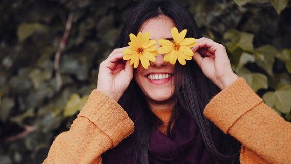 Eine lachende junge Frau steht vor einer Hecke und hält sich Blumen vor die Augen. © Manu Reyes Foto: Manu Reyes