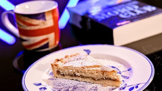 Eine Stück Bakewell Tart neben einem Becher mit britischer Fahne un dienem Buch - Folge 64  von eat.READ.sleep © NDR Foto: Claudius Hinzmann