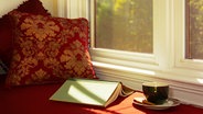 Ein aufgeschlagenes Buch liegt auf einer Fensterbank neben einer Kaffeetasse. © iStock Foto: Diane Diederich
