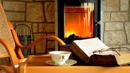 Ein aufgeschlagenes Buch liegt neben einer Kaffeetasse vor einem Ofen. © Fotolia Foto: K.-U. Häßler