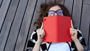Eine Frau, die auf einem Holzsteg liegt, hält sich ein Buch vor Mund, Hals und Nase. © fotolia.com Foto: pintorama