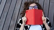 Eine Frau, die auf einem Holzsteg liegt, hält sich ein Buch vor Mund, Hals und Nase. © fotolia.com Foto: pintorama