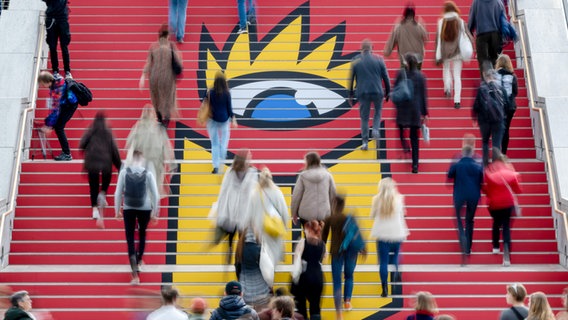 Besucher der Leipziger Buchmesse gehen zur Eröffnung über eine Treppe mit dem Logo der Buchmesse. © picture alliance/dpa | Hendrik Schmidt 