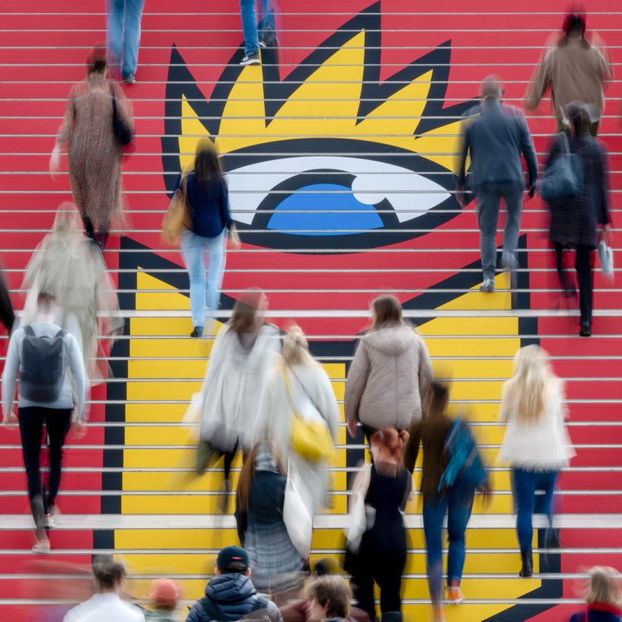 Besucher der Leipziger Buchmesse gehen zur Eröffnung über eine Treppe mit dem Logo der Buchmesse. © picture alliance/dpa | Hendrik Schmidt 