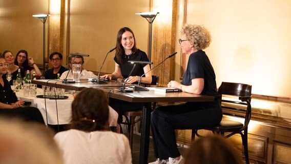 Eine Frau mit breitem Lächeln wird von einer anderen Frau vor einem Publikum interviewt Eine Frau mit breitem Lächeln wird von einer anderen Frau vor einem Publikum interviewt © Literaturhaus Hamburg Foto: Daniel Müller