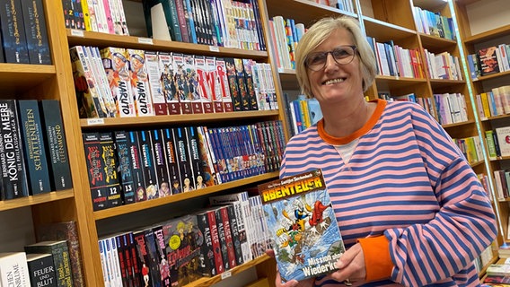 Christiane Hoffmeister steht mit einem "Lustigen Taschenbuch" vor dem Comic- und Manga-Regal ihrer Buchhandlung © NDR.de Foto: Mathias Heller