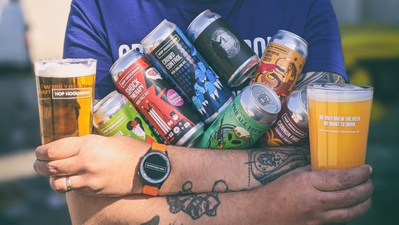 Eine Person hält sieben verschiedene, bunte Bierdosen der Marke Hop Hooligan in den Armen, in jeder Hand ein gefülltes Glas derselben Marke (Aufschrift: "We only brew the beer we want to drink) © Design Diana Barbu, Photo Courtesy of Hop Hooligans, Craft Beer Design, gestalten 2022 