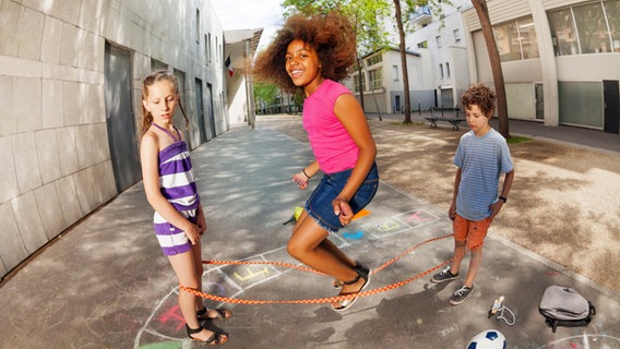 Drei Kinder spielen Gummitwist auf einem Schulhof. © IMAGO / agefotostock 
