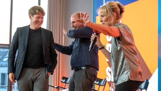 Zwei Männer und eine Frau auf der Bühne lachen und winken mit Mikros - die Hosts des Literaturpodcasts eat.READ.sleep.
im Literaturhaus München © NDR / Catherina Hess Foto: Catherina Hess