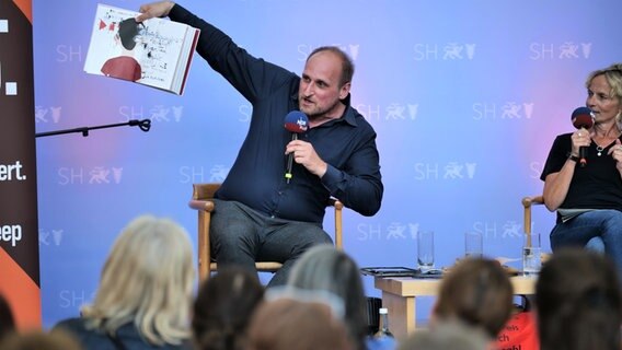 Host Daniel Kaiser hält bei einer Podcastsession in Berlin ein Buch hoch © NDR Foto: Torsten Heitmann