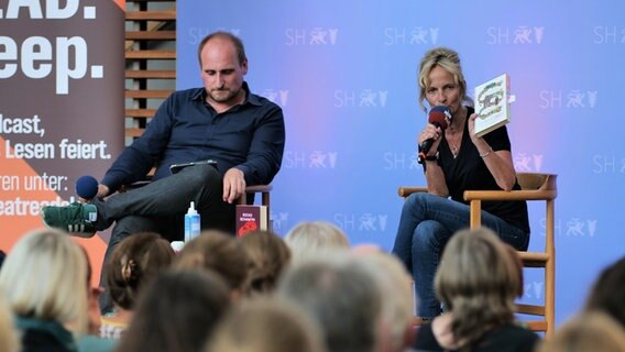 Zwei Personen auf einer Bühne sitzen vor Publikum - Host Katharina (rechts) hält ein Buch hoch © NDR Foto: Torsten Heitmann