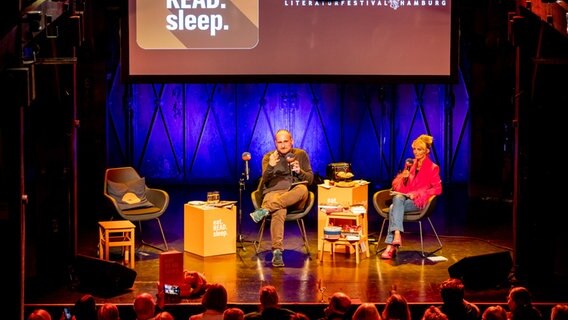 Ein Mann und eine Frau sitzen auf einer Bühne -Daniel Kaiser und Katharina Mahrenholtz moderieren live eine Podcastfolge eat.READ.sleep beim Harbour Front Literaturfestival in Hamburg © NDR Foto: Philipp Szyza