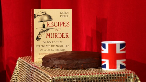 Ein Kuchen mit Schokoladenkovertüre neben einem Becher mit Großbritannienflagge und einem Buch -  Folgenfoto von eat.READ.sleep © NDR Foto: Katharina Mahrenholtz