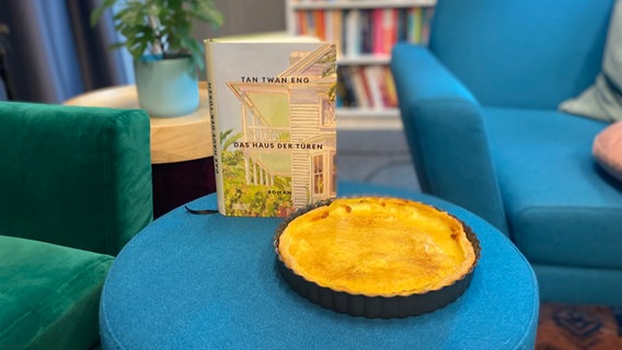Ein Buch steht auf einem blauen Hocker, daneben steht eine runde Backform mit etwas flachem braunen Gebäck wie eine Quiche © NDR Foto: Katharina Mahrenholtz
