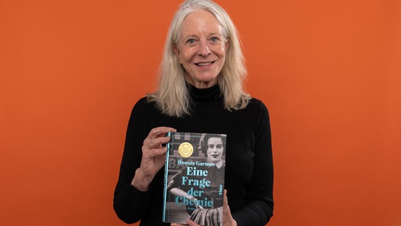 Eine Frau mit weißen Haaren schaut lächelnd in die Kamera und hält ein Buch hoch - Bonnie Garmus © NDR / Ralf Plessmann Foto: Ralf Plessmann
