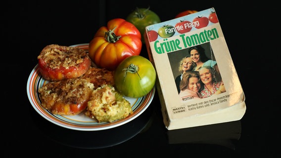 Auf schwarzem Hintergrund ist ein Teller mit roten, grünen und aufgeschnittenen, überbackenen Tomaten angerichtet. Daneben liegt Fanny Flaggs Roman "Grüne Tomaten", auf dem Titelbild die Protagonistinnen der Verfilmung, darunter die Oscarpreisträgerinnen Kathy Bates und Jessica Tandy. © NDR Foto: Ekaterina Shurygina