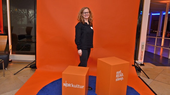 Eine Frau mit Locken, dunkler Brille steht bei einer orangefarbenen Fotoecke zum Fanfest der 100. Folge von eat.READ.sleep in Hannover © NDR Foto: Patricia Batlle