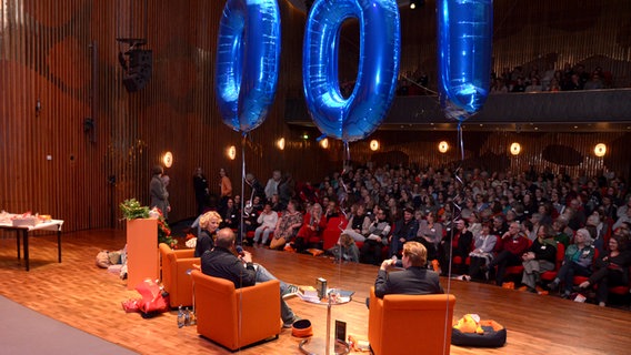 Blaue Ballons mit der Ziffer 100 schweben über drei Menschen auf Sesseln im Konzertsaal des NDR in Hannover, das Publikum lauscht der Veranstaltung © NDR Foto: Patricia Batlle