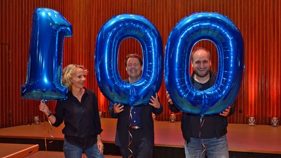 Drei Podcasthosts schauen an blauen Luftballos vorbei und lächeln - die Luftballons bilden die Ziffer "100" © NDR Foto: Patricia Batlle