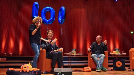 Eine Frau steht und lächelt, zwei Männer sitzen in orangefarbenen Sesseln - Die Host Katharina, Jan und Daniel (von links) beim Fanfest zur 100. Folge von eat.READ.sleep in Hannover © NDR Foto: Patricia Batlle