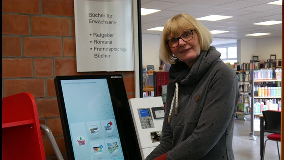 Eine Frau hält Bücher steht neben einem Selbstverbuchungsautomaten - im Hintergrund Bücher © Helgard Füchsel Foto: Helgard Füchsel