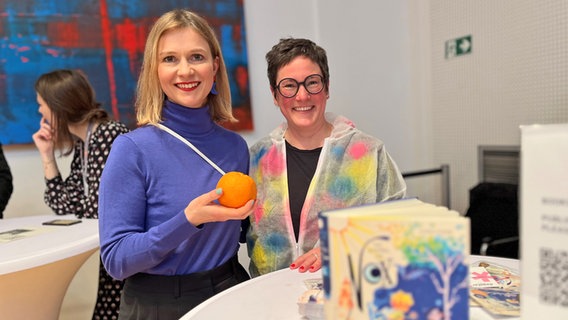 Eine Frau mit blauen Pulli ält eine Orange, danaben seht eine Frau mit dunkler Brille (Autorin Claudia Scharf mit Agentin Danile Steiner von Carlsen bei "Books at Berlinale") © NDR Foto: Patricia Batlle