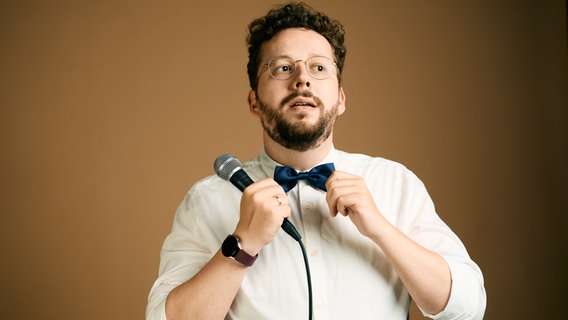 ein Mann mit hellem Hemd, braunen Locken und Brille hält sich eine Fliege und ein Mikro in der Hand (Der Autor Henrik Szántó) © Marvin Ruppert Foto: Marvin Ruppert