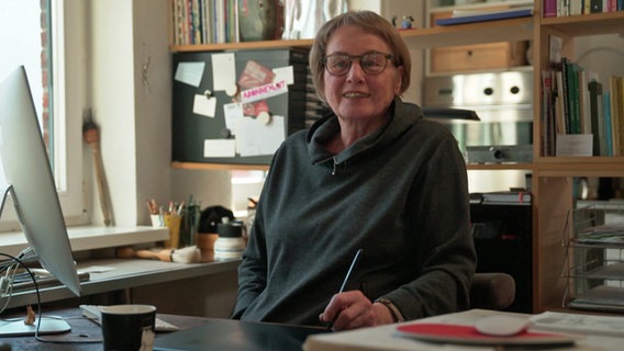 Die Illustratorin Jutta Bauer sitzt in einem dunklen Hoody an ihrem Schreibtisch. © NDR 