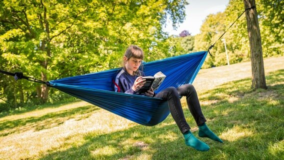 Eine junge Frau liest ein Buch in einer Hängematte, die zwischen zwei Bäumen gespannt ist. © picture alliance/dpa | Moritz Frankenberg 