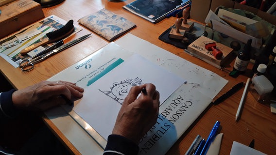 Die Hände von Paul Maar in Nahaufnahme - in der Rechten ein Stift, mit dem er gerade ein Sams auf ein Blatt Papier zeichnet. © NDR Foto: Katja Weise