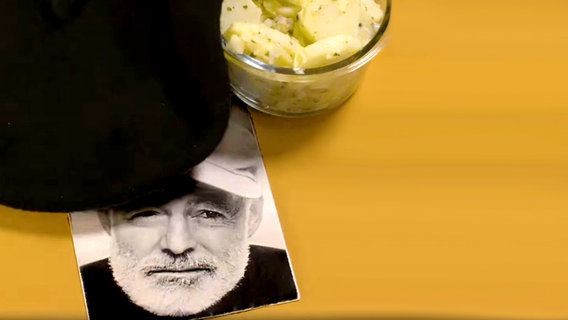 Eine Schale Kartoffelsalat mit einer Baskenmütze und einem Bild von Ernest Hemmingway. © NDR Foto: Katharina Mahrenholtz