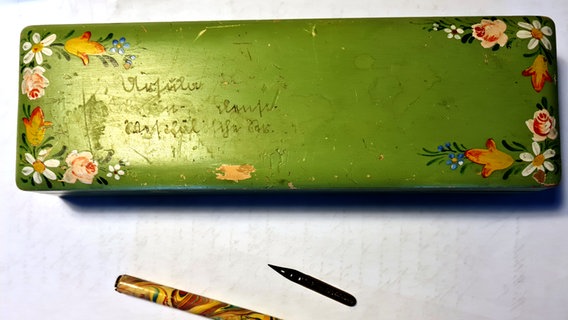 Ein grüner Schreibkasten, darauf eine Inschrift in Sütterlinschrift, darunter ein Blatt Papier und eine Feder © NDR Foto: Agnes Bührig