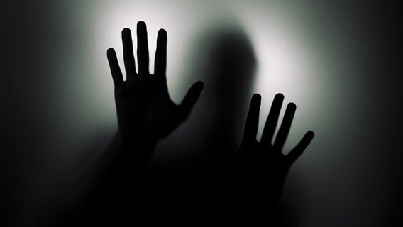 Dunkle Silhouette mit vorgehaltenen Handflächen hinter einer Glasscheibe. © fotolia.com Foto: Jakub Krechowicz