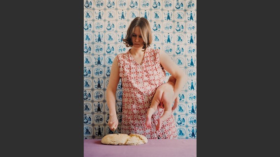Foto aus dem Bildband "Mama": Eine Frau schneidet Brot und hält in der anderen Hand ihr Baby © Stiftung Museum Kunstpalast, Düsseldorf Foto: Judith Samen