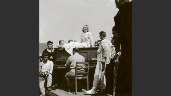 Foto aus dem Bildband "Marlene Dietrich an der Front": Schwarzweiß-Foto von einer blonden Frau, die umringt von Soldaten auf einem Klavier sitzt und singt. © Deutsche Kinemathek - Marlene Dietrich Collection Berlin 