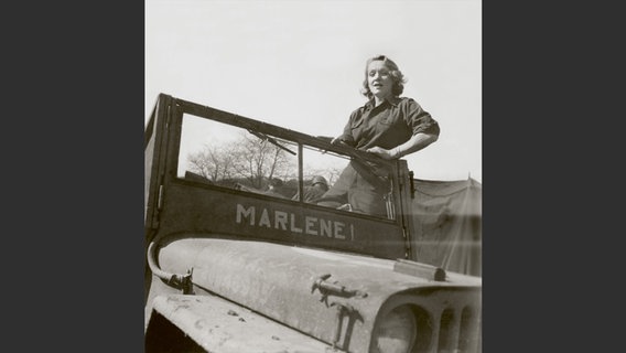Foto aus dem Bildband "Marlene Dietrich an der Front": Schwarzweiß-Foto von einer blonden Frau, die in einem Militär-Jeep steht. © Deutsche Kinemathek - Marlene Dietrich Collection Berlin 