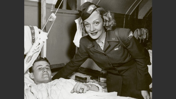 Foto aus dem Bildband "Marlene Dietrich an der Front": Schwarzweiß-Foto von einer blonden Frau in Militäruniform, die am Bett eines Verwundeten steht. © Deutsche Kinemathek - Marlene Dietrich Collection Berlin 