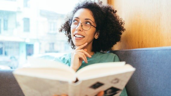 Eine Frau hat ein aufgeschlagenes Buch in der Hand und guckt nachdenklich nach oben. © picture alliance / Westend61 | alev 