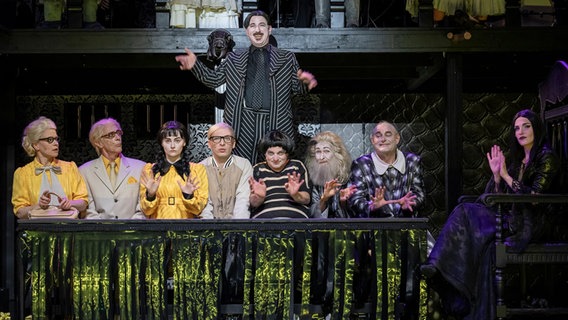 Mitglieder einer gruseligen Familie hinter einem Tisch auf einer Theaterbühne schauen ins Publikum. Mitglieder einer gruseligen Familie hinter einem Tisch auf einer Theaterbühne schauen ins Publikum. © Volker Beinhorn Foto: Volker Beinhorn