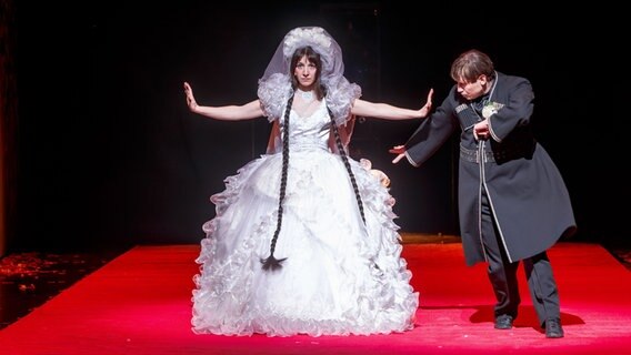 Schauspielerin Rosa Thormeyer steht im Hochzeitskleid auf der Bühne und streckt die Arme aus. Ole Lagerpusch tanzt um sie herum. © picture alliance/dpa | Markus Scholz 