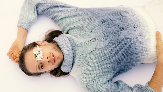 Ein Mann mit hellblauem Strickpulli liegt auf dem Rücken und hat eine weiße Blume auf einem Auge. © Schauspiel Hannover/ Kerstin Schomburg Foto: Kerstin Schomburg