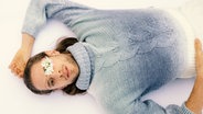 A man in a light blue knitted sweater is lying on his back and has a white flower in one eye.  © Schauspiel Hannover / Kerstin Schomburg Photo: Kerstin Schomburg