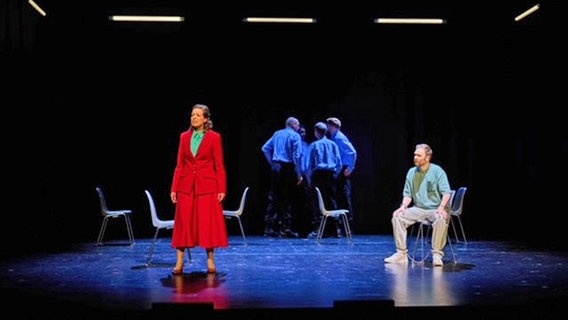 eine Frau im roten Kostüm mit grüner Bluse steht auf der Bühne, dahinter stehen mehrere Männer im Hintergrund, im Vordergrund rechts sitzt ein Mann mit Fußfesseln © SH Landestheater / Thore Nilsson Foto: Thore Nilsson