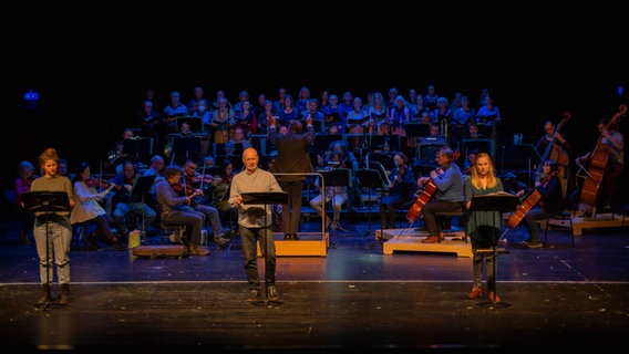 Drei Schauspieler stehen auf der Bühne vor einem Orchester und Chor © Theater Lüneburg/ joeigraphie Foto: joeigraphie