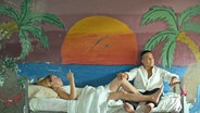 Eine Frau liegt in einem Gefängnisbett, ist zugedeckt und raucht, eine andere sitzt daneben, dahinter ist eine bunte Palmenlandschaft an die Wand gemalt - Szenebild aus der Theaterproduktion "Lola Arias Reas" von Lucinda Childs auf Kampnagel © Camilla Greenwell, Belinda Lowley Foto: Camilla Greenwell, Belinda Lowley